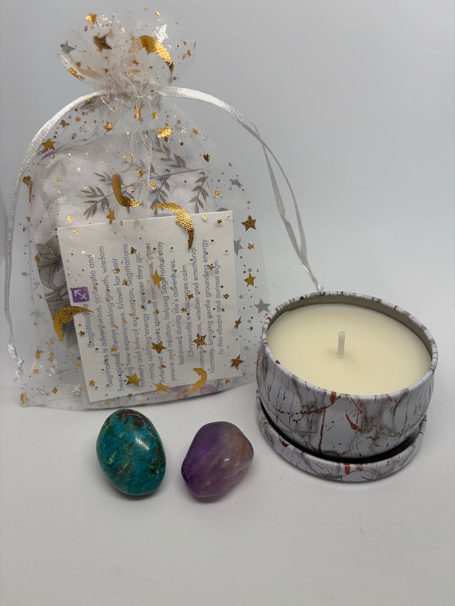 Candle in a decorative tin with two stones and a clear bag with text on a light gray background